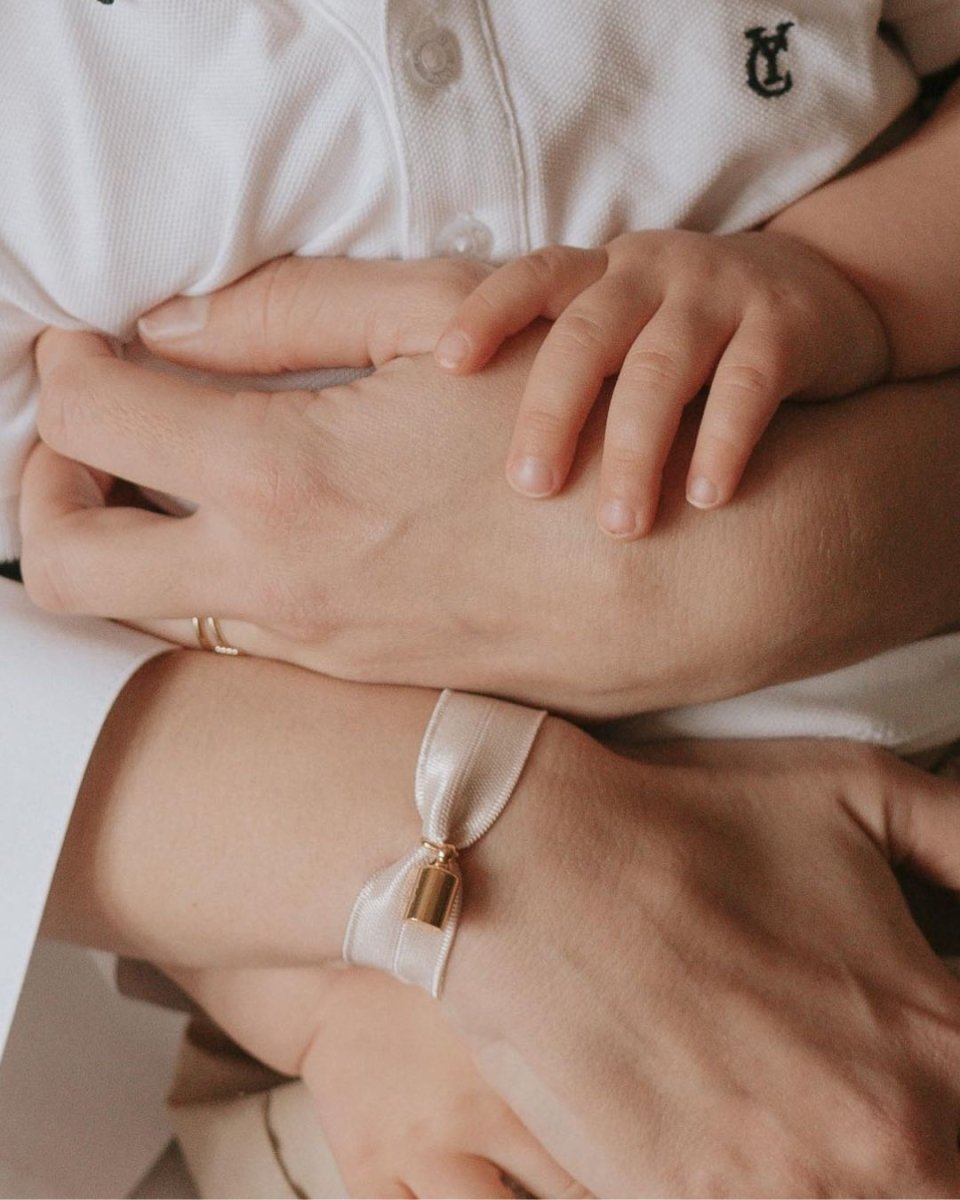 Pendentif Mon Petit Poids plaqué or jaune sur un ruban irisé nude, autour du poignet d'une femme, mains entrelacées celles d'un enfant.