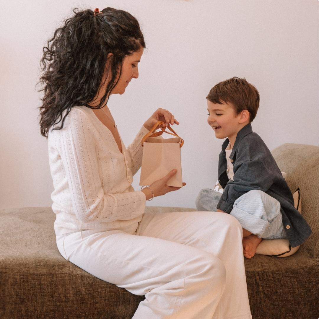 Une maman tient un sac cadeau dans ses mains et son fils le regarde en souriant.