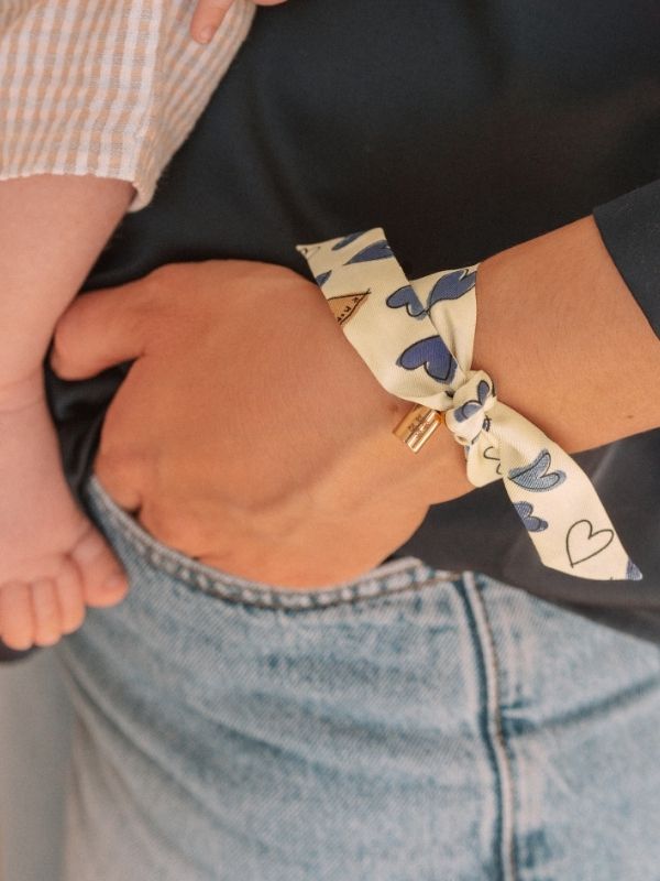 Bracelet en soie imprimée bleu et beige avec pendentif personnalisé en plaqué or jaune, porté au poignet avec un pied de bébé en arrière-plan