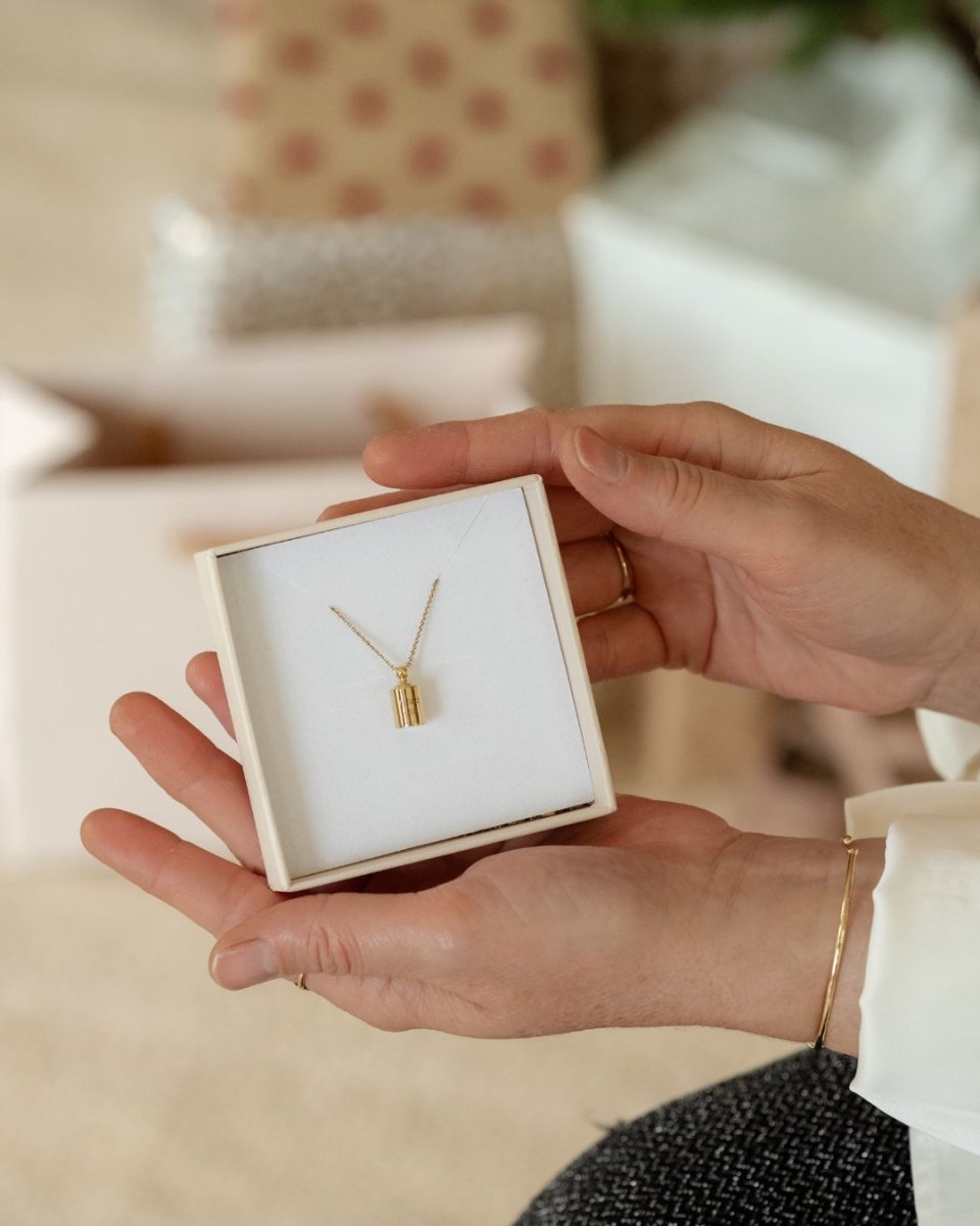 Collier Mon Petit Poids plaqué or jaune dans son écrin, tenu par les mains d'une femme - fond flou