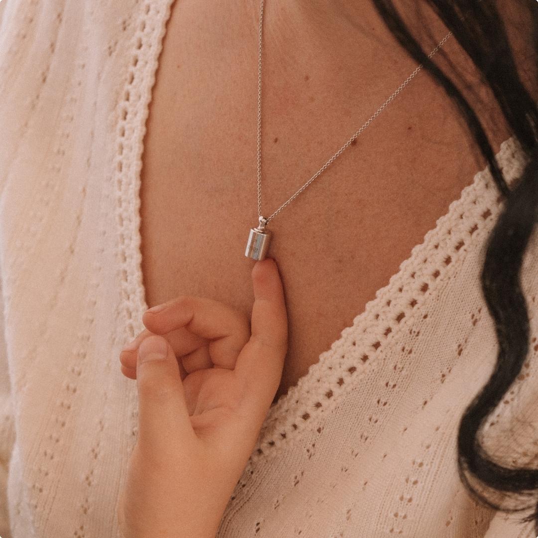 Collier Mon Petit Poids argenté rhodié porté au cou d'une femme avec une main d'enfant qui le touche du bout du doigt.
