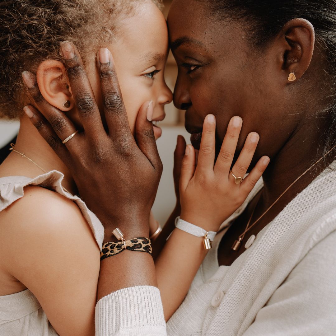 Une maman et sa fille se regardant dans les yeux, front contre front, se tenant les joues. Elles portent des bracelets Mon Petit Poids. 