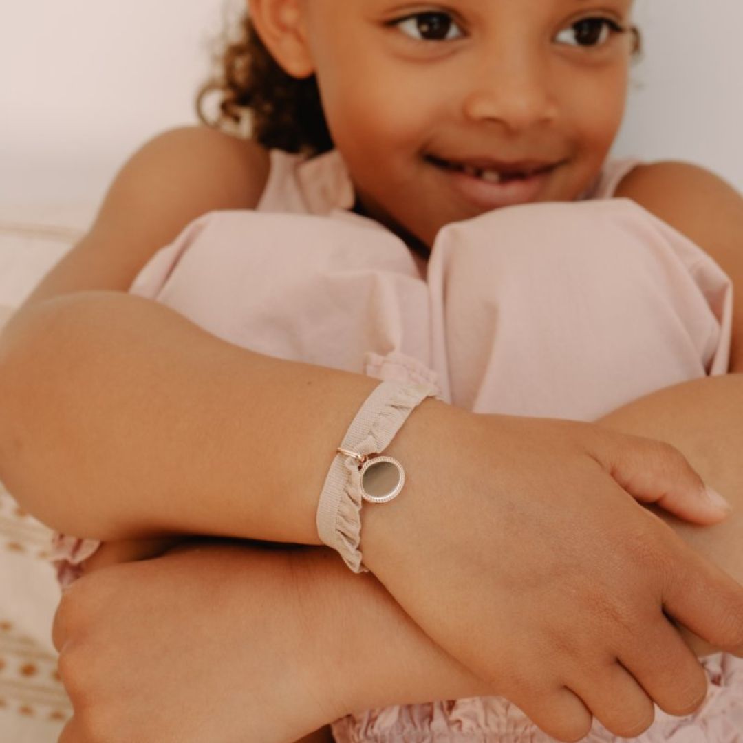 Bracelet Médaille Madeleine avec un ruban frou rose poudré, attaché autour du poignet d'une petite fille qui a les bras autour des genoux