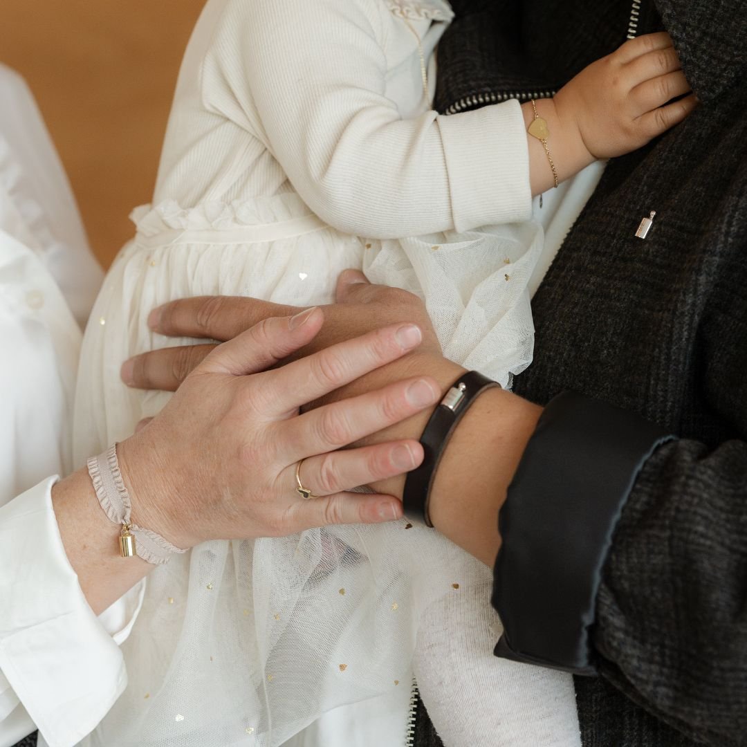 Mains portant un bébé. Chacune est habillé d'un bracelet. A gauche, un braceletMon petit Poids avec un froufrou rose poudré. A droite un bracelet en cuir brun